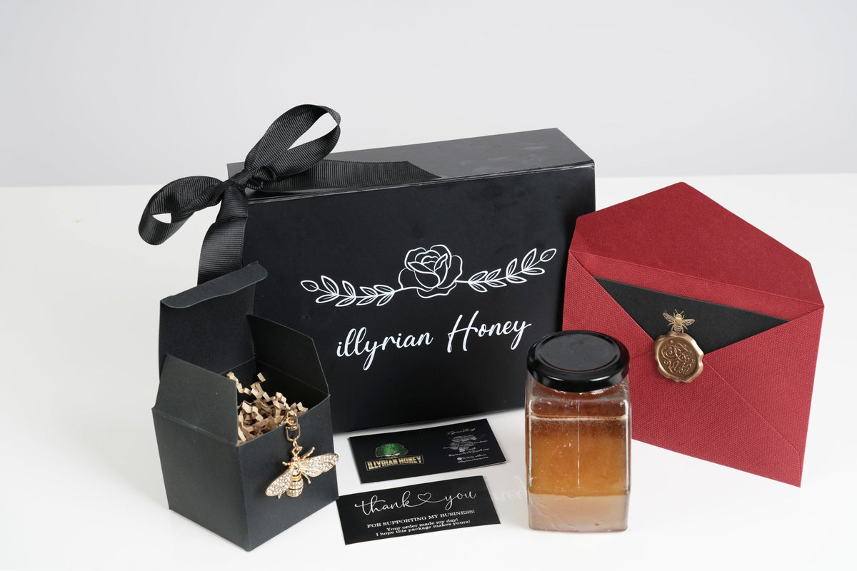 A premium honey gift set by Illyrian Honey, featuring a black presentation box with a black ribbon, an open box showing a gold beehive necklace, a glass jar of honey, a red envelope with a gold wax seal, and business cards on a white background.